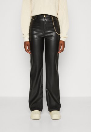 Person iført sorte bukser i kunstlæder med guldkæde, cremefarvet strikket sweater og offwhite sko står mod en simpel baggrund.