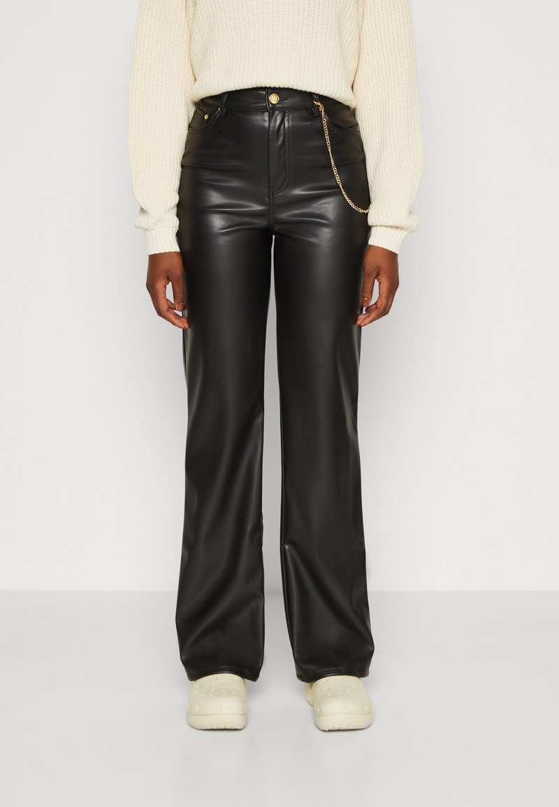 Personne portant un pantalon noir en simili cuir avec une chaîne dorée, un pull en maille crème et des chaussures écrues, debout devant un fond uni.