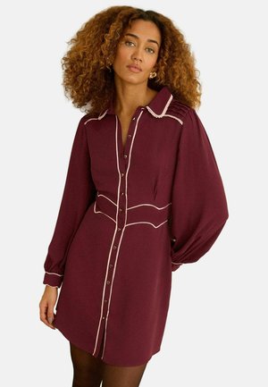 Kvinde med krøllet hår iført en bourgognefarvet kjole med knapper, hvid piping, lange puffede ærmer og gyldne creoler.