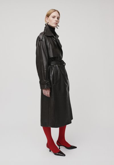 Eine Frau in einem schwarzen Leder-Trenchcoat, einem schwarzen Rollkragenpullover, roten Strumpfhosen, schwarzen spitzen Absätzen und auffälligen Ohrringen steht vor einem schlichten Hintergrund.