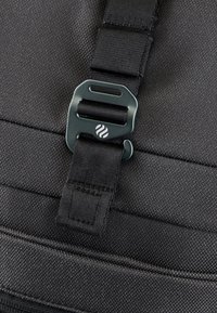 Sangle en tissu noir avec une boucle de fermeture sombre ; matériau texturé avec un motif en grille et un petit logo sur la boucle.