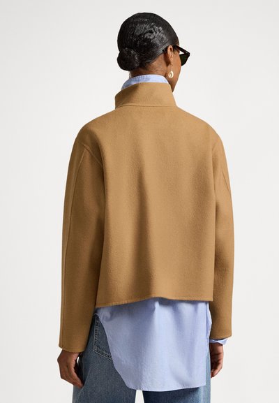 Καφέ μπουφάν από φλις με ψηλό γιακά και σχεδίαση cropped, φορεμένο πάνω από ένα ανοιχτό μπλε πουκάμισο. Διαθέτει καθαρή, απλή σιλουέτα.
