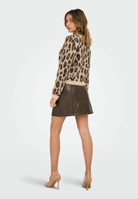 Žena u bež i smešim leopard printu svetrima, tamnosmeđoj kožnoj suknji i bež cipelama na visoku petu, stoji i gleda unazad.