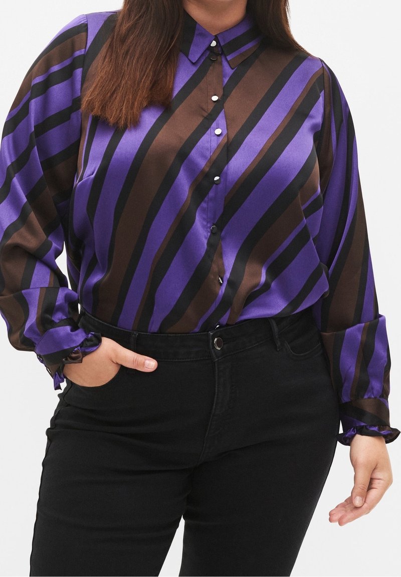 Personne portant un chemisier à boutons à rayures diagonales violet, marron et noir avec des poignets volantés et un pantalon noir taille haute, main dans la poche.