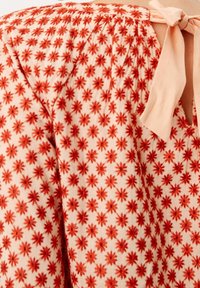 Blomstret bluse med et let ferskenfarvet stof og røde blomstermønstre, smock-detaljer ved skuldrene og en snørelukning bagpå.