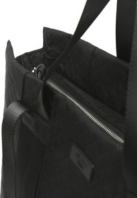 Sac en tissu matelassé noir avec une poche zippée et des poignées lisses. L'intérieur présente une doublure en nylon et un détail de marque subtil.