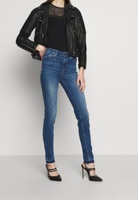 Svart läderbikerjacka med ribbade detaljer, svart topp med spetstillbehör, blå skinny jeans och svarta spetiga klackar.