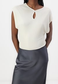 Bezrękawnik w kolorze kremowym z dekoltem w kształcie dziurki, połączony z ciemnoszarym satynowym kobiecym spódnicą o wysokim stanie, na osobie z rękami za plecami.