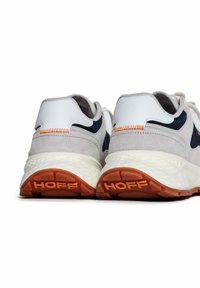 Baskets de course blanches et grises en matériaux en daim et cuir, avec des accents orange et des semelles texturées. Nom de la marque "HOFF" au niveau du talon.