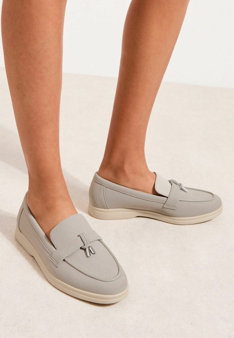 Mocassins gris clair à enfiler avec semelles blanches basses et pompons décoratifs, portés pieds nus sur un fond neutre.