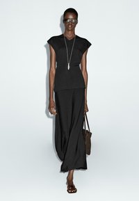 Top nero a maniche corte e gonna lunga e fluida di colore nero realizzata in un tessuto leggero. La modella indossa occhiali da sole scuri e porta una borsa marrone.