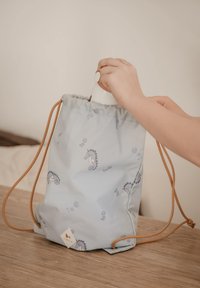 Vaaleansininen nyörilaukku merihevospainatuksella. Valmistettu pehmeästä kankaasta, jossa ruskeat naruhihnat ja kolmionmuotoinen logotunniste.