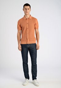 Pfirsichfarbener, gestrickter Polo mit kurzen Ärmeln und einem strukturierten Kabelmuster, ausgestattet mit einem Kragen und fünf Knöpfen, kombiniert mit dunkelblauen Jeans und weißen Turnschuhen.