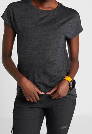 Osoba w ciemnej koszulce z krótkim rękawem i ciemnych spodniach, nosząca smartwatch z żółtym paskiem, poprawia koszulkę przy talii.