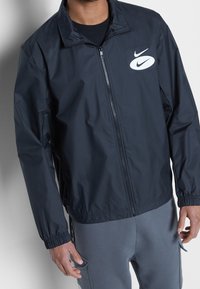 Man in een marineblauwe zip-up jack met elastische manchetten en witte Nike-logo's op de borst, gecombineerd met grijze broek.