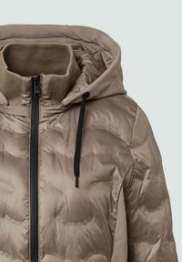 Chaqueta acolchada beige con cierre de cremallera, capucha y cuello acanalado. La textura es acolchada y cuenta con cordones negros para ajuste.