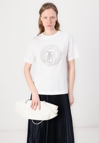 Hvit bomulls t-skjorte med et rundt grafisk design i sølv i midten, sammen med en hvit plissert skjørt og en hvit lær håndveske.
