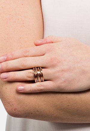 Set di anelli in oro rosa con bande intrecciate, texture liscia e finitura lucida. Indossati su dita contro un braccio di colore chiaro.