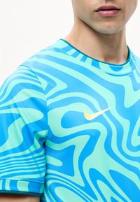 Κοντινό πλάνο ενός ατόμου που φοράει μπλε και τυρκουάζ μπλουζάκι Nike με κυματιστό σχέδιο και κίτρινο λογότυπο Swoosh στο στήθος.