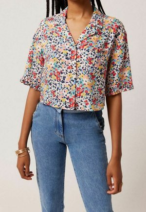 Kvinde iført en kortærmet, flerfarvet blomstret knappeskjorte med krave og højtaljede blå jeans.