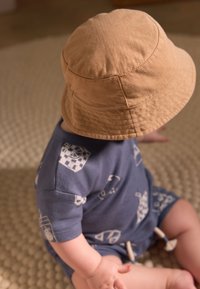 Chapeau seau marron clair sur un enfant, assorti à une tenue bleue avec des motifs géométriques blancs. Textures douces et coupe décontractée.