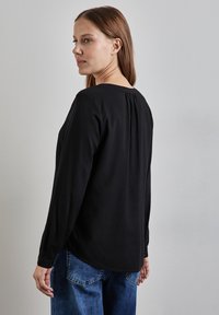 Chemise noire à manches longues avec une coupe décontractée, dotée d'un col rond et de détails plissés au dos, associée à un jean en denim bleu.