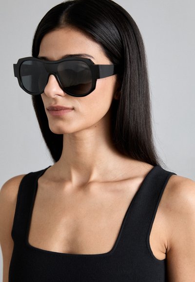 Lunettes de soleil surdimensionnées noires avec une monture épaisse, un design angulaire et des verres sombres dégradés ; portées avec un haut sans manches noir.