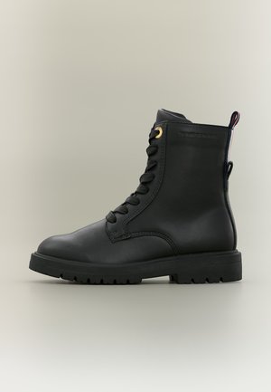 Bottine noire en cuir à lacets avec semelle épaisse et petite languette en tissu à l’arrière sur un fond neutre.