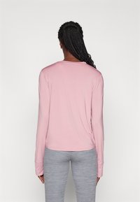 Long-sleeve shirt in light pink, made of soft fabric. Features a relaxed fit with rounded hem and wrist cuffs. Paired with gray leggings.