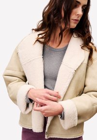 Vrouw met golvend bruin haar in een beige schapenvachtjas over een grijs shirt, met haar handen dicht bij haar taille.