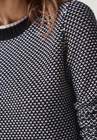 Czarno-biały sweter dziany z wzorem serca, z prążkowanym kołnierzem i teksturowaną tkaniną, zbliżenie na rękaw i okolicę ramienia.