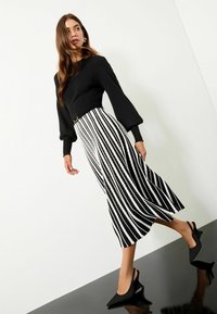 Top negru cu dungi, cu mâneci voluminoase, purtat cu o fustă mid-length cu dungi albe și negre și pantofi cu toc ascuțit, din material textil negru, cu slingback.