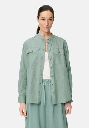 Femme aux cheveux foncés attachés en queue de cheval, portant une chemise boutonnée vert clair sur un haut blanc et un pantalon vert clair assorti à cordon de serrage.