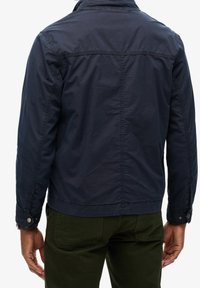 Granatowa lekka kurtka z kołnierzem stójką, widocznymi przeszyciami, zapinanymi na guziki mankietami i prostym krojem w talii.