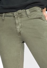 Khaki spalvos liesos džinsai su lygiu paviršiumi, paspaudimo uždarymu, siuvinėtomis detalėmis ir prigludusia išvaizda.