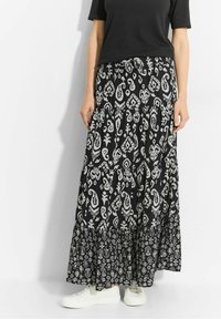 Jupe longue noire avec un motif blanc complexe ; design à étages, tissu doux et ourlet volanté. Associée à des baskets blanches pour le contraste.