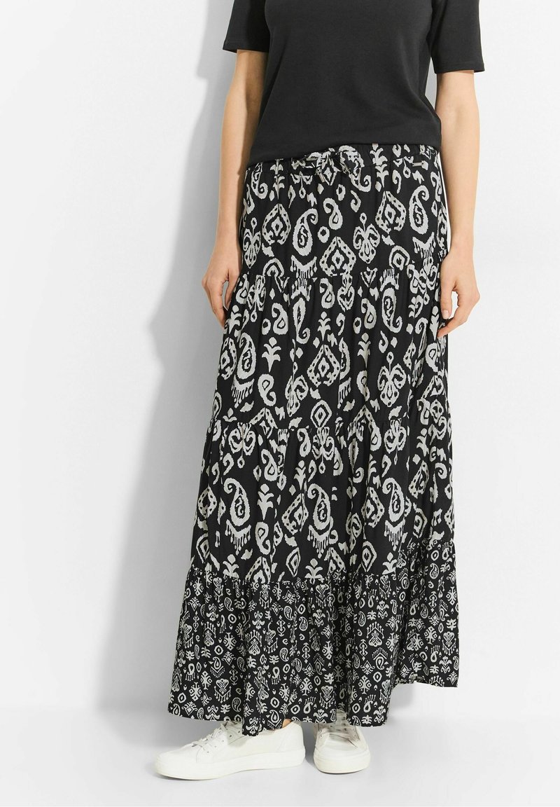 Jupe longue noire avec un motif blanc complexe ; design à étages, tissu doux et ourlet volanté. Associée à des baskets blanches pour le contraste.