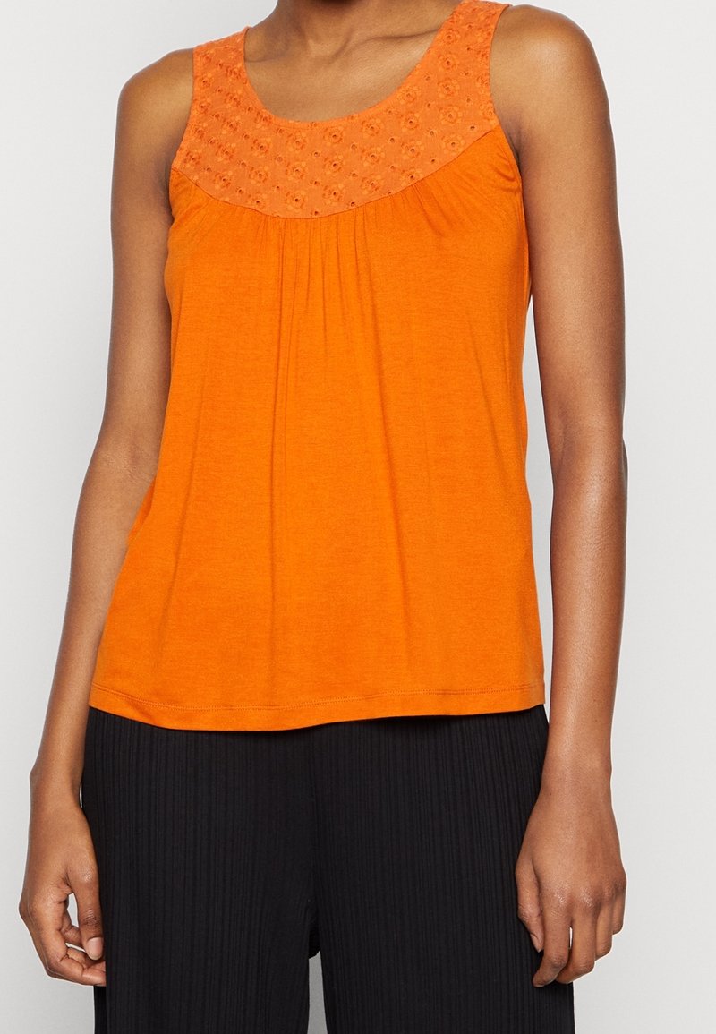 Haut sans manches orange vif avec un empiècement brodé floral, porté avec un pantalon noir côtelé, visible des épaules aux hanches.