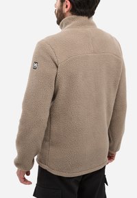 Homme portant une veste en polaire beige avec un logo sur la manche gauche, vu de dos, associé à un pantalon cargo noir.