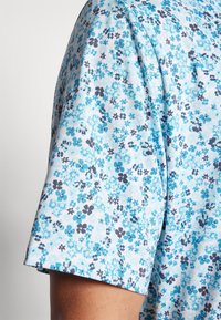Chemise à manches courtes en tissu bleu clair avec un motif floral comprenant des nuances de bleu et de gris foncé, surface texturée et finition douce.