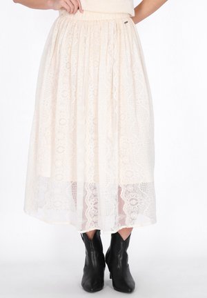 Femme portant une jupe midi en dentelle crème avec une ceinture élastique et des bottines noires, debout devant un fond blanc.