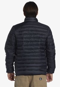 Marinblå puffjacka med quiltat mönster, hög krage och elastiska ärmslut. Tillverkad av ett glänsande, lätt material.