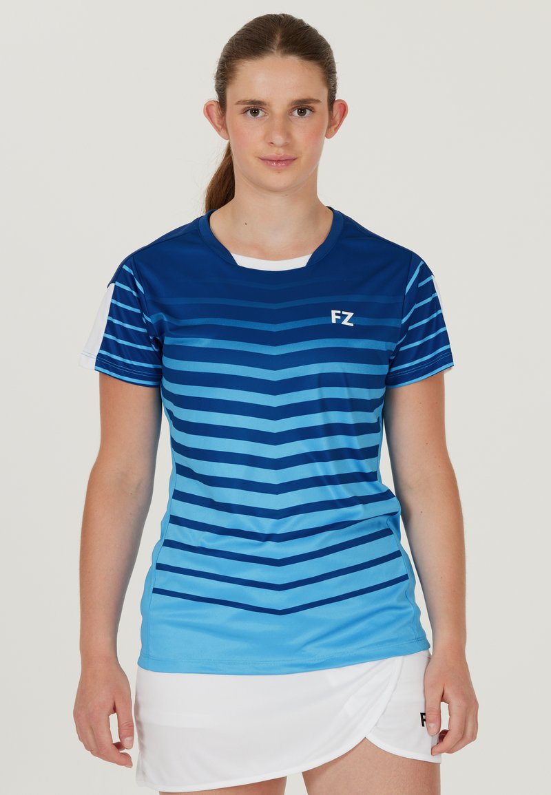 Ung kvinna med brunt hår i hästsvans, iklädd blå randig sporttröja och vit kjol, stående mot en enkel bakgrund.