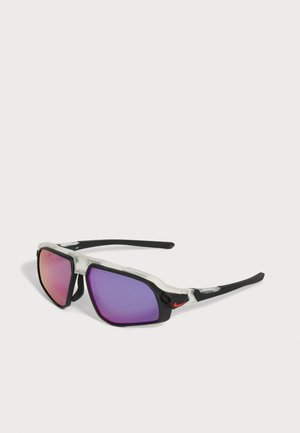 Gafas de sol deportivas con montura negra y translúcida, lentes espejadas moradas y un pequeño logo rojo de swoosh en la patilla.