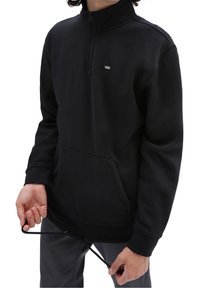 Uomo che indossa una felpa nera Vans con zip a quarto e tasca a marsupio, mentre regola il cordino all'orlo, su uno sfondo bianco.