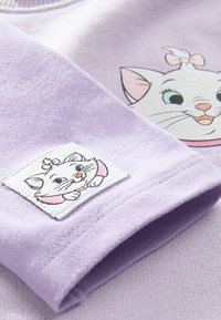 Lila polár pulóver, amelyen mese macska mintázat és hímzett folt található az ujjon. Puha tapintású, bordázott gallérral és aljjal.