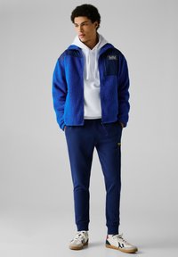 Fleecejacke in leuchtendem Blau mit einem dunkelblauen Einsatz, weißem Hoodie darunter, marinefarbene Jogginghose und weißen Sneakern mit kontrastierenden Akzenten.