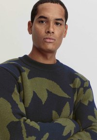 Strickpullover mit dunkelblauem Grund, großflächigen grünen Blattmustern, rundem Ausschnitt und gerippten Bündchen. Weicher Griff, lässiger Schnitt.