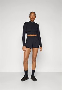 Veste noire courte zippée à manches longues associée à un short plissé noir. Le mannequin porte des chaussettes et des chaussures noires, debout sur un fond neutre.