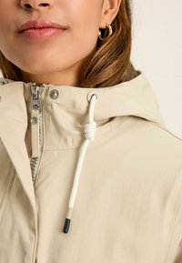 Jacket cu glugă de un bej deschis, cu fermoar frontal, șnur cu accent legat și detalii de metal pe guler.